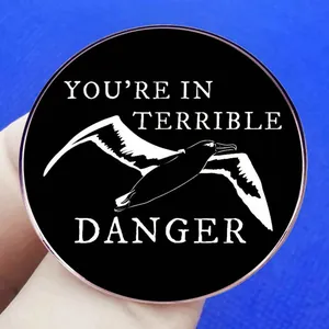 Black round seagull enamel pin with 'You're in Terrible Danger' text held by hand