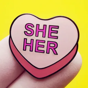 Spilla in smalto rosa a forma di cuore con scritta She Her su sfondo giallo