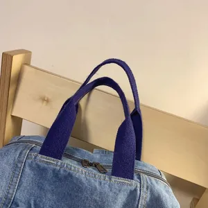 Detail of light blue denim handbag with purple handles and dual metal zippers on wood chair