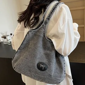 Washed dark gray denim tote bag with metal bow emblem shown on model in a cafe setting