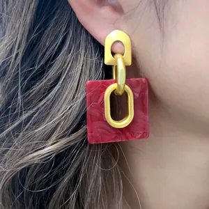 Detalle de pendientes geométricos cuadrados con acabado dorado mate y resina roja