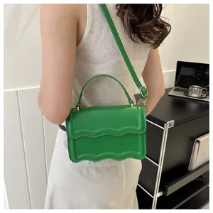 Green square crossbody bag with wavy flap shown on shoulder in a modern indoor setting