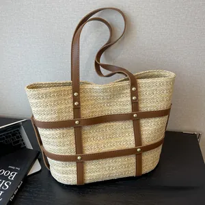 Minimalist straw tote bag with brown leather straps and gold stud details on gray backdrop
