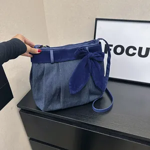 Blue denim crossbody bag with navy velvet bow and pleated design on black wood surface