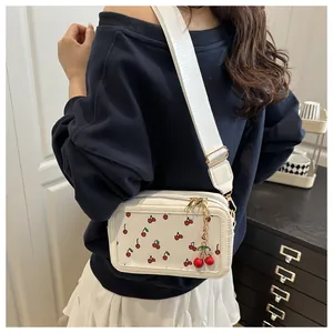 Close-up of white crossbody bag with cherry print, wide strap and cherry pendant charm