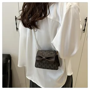 Detail of dark brown square bag with M-pattern, bow accent, and pearl shoulder strap