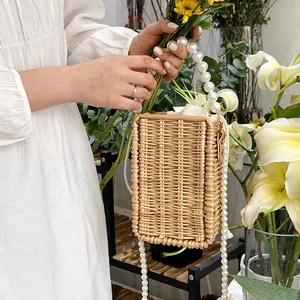 Handwoven rattan box bag featuring pearl handles and strap against a floral backdrop