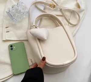 Dettaglio borsa a secchiello in pelle crema con ciondolo a cuore accanto a un cellulare