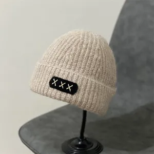 Detail of beige mohair knit beanie featuring a black square patch with white X stitching