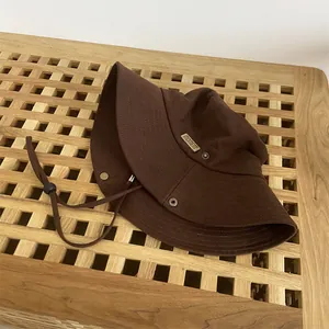 Detail of a brown canvas bucket hat featuring a metal 'MOMENT' logo and adjustable drawstring