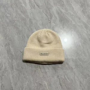 Dettaglio berretto in maglia a coste beige con targhetta in metallo argento incisa