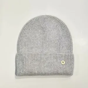 Detalle de gorro de punto gris claro con textura suave y etiqueta metálica con logo 'B'