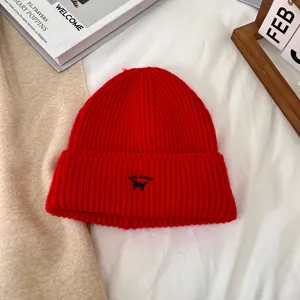 Detalle de gorro de punto rojo con bordado de gato azul sobre ropa de cama blanca