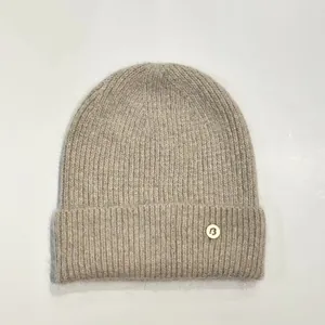 Detalle de un gorro de punto acanalado beige con un emblema metálico letra B en la vuelta