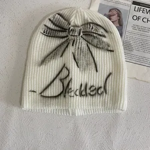 White ribbed knit beanie with black graffiti bow and Blessed text on cream background