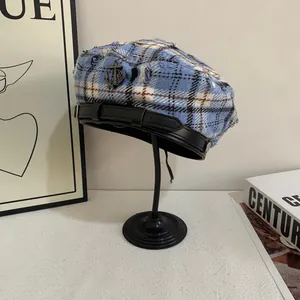 Detail van de blauw geruite tweed baret met metalen ankerembleem en zwart lederen band