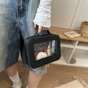 Black rectangular cosmetic bag with transparent window and gold zippers held by a hand