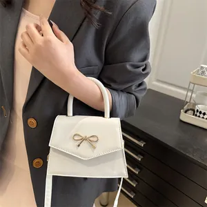 White minimalist crossbody bag with gold bow clasp shown held by a model in a suit