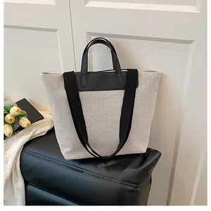 Minimalist canvas tote bag with black leather trim and wide shoulder strap on a quilted bench