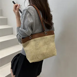 Straw woven tote bag with brown faux leather trim shown on shoulder in a minimalist setting
