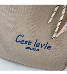 Close-up of the beige canvas tote bag with deep blue C'est lavie MDL/BAG embroidery detail