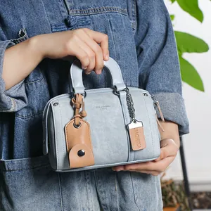 Detail view of light blue denim Boston bag with tan leather trim and metal chain charm