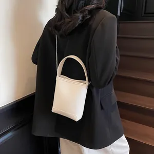 Minimalistische beige emmertas van leer met handvat en een schouderriem van metalen ketting