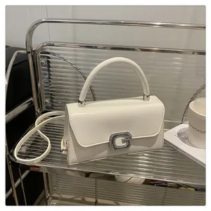 Close-up of white crossbody bag featuring a silver rhinestone G-buckle on transparent shelf