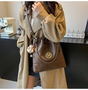Détail du sac seau en cuir marron foncé avec logo M doré, chaîne et breloque peluche