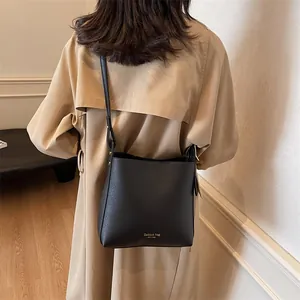 Minimalist black leather bucket bag with wide strap worn by model in beige trench coat