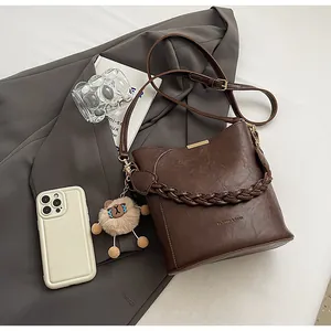 Detail einer braunen Eimer-Tasche mit gewebtem Griff, Text und kleinem Bären-Anhänger
