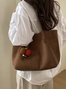 Brown faux suede tote bag with decorative pompom charms worn over a woman's shoulder