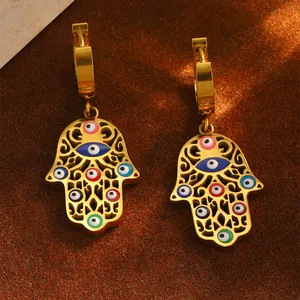 Detailansicht goldener Hamsa-Hand-Ohrringe mit bunten Evil-Eye-Anhängern auf braunem Stoff