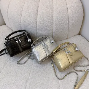 Tres bolsos caja con patrón de cocodrilo en negro, plata y oro sobre sofá de felpa blanca