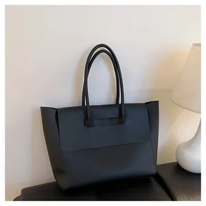 Minimalist black leather tote bag with flap closure placed on a soft black cushion