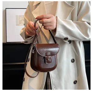 Dettaglio borsa a sella marrone scuro lucida indossata sopra un trench minimalista