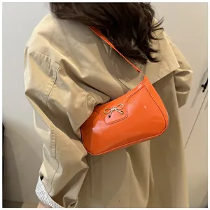 Detail of glossy orange PU leather baguette bag featuring a gold bow accent on a model
