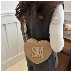Detail of heart-shaped khaki crossbody bag with SM logo and wide woven strap on a model