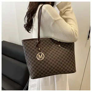 Detail of dark brown monogram tote bag featuring a gold M-shaped metal pendant and leather handles