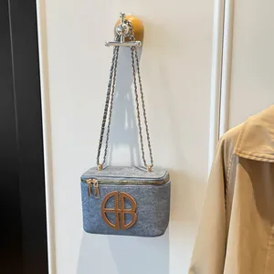 Sac bandoulière en feutre gris avec chaîne dorée et logo suspendu sur un mur blanc