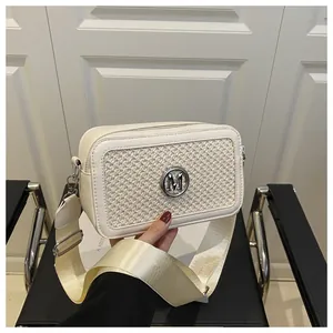 Detail of a woven cream crossbody bag featuring a metallic M logo and wide nylon strap