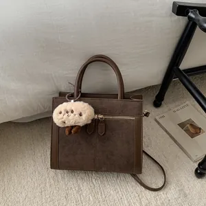 Bolso tote de ante marrón con detalles de cuero y colgante de peluche sobre una alfombra