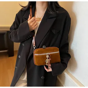Detail of a brown suede crossbody bag featuring a silver zipper and a robot charm on a model