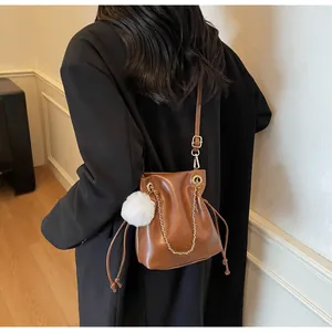 Détail d'un sac seau en cuir marron avec chaîne dorée, pompon blanc et fermeture à cordon