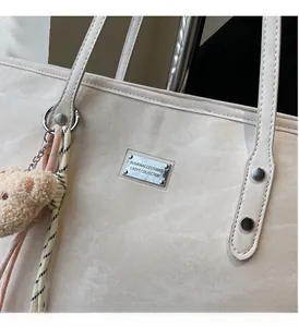 Detail of a creamy white tote bag featuring a silver brand plate and plush bear charm