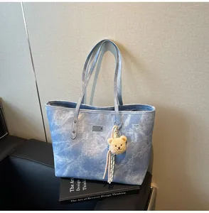 Detail of light blue denim tote bag with plush bear charm on black leather sofa
