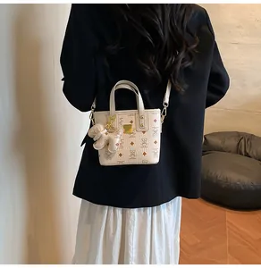Detail of cream printed tote bag with bear charm and golden logo plate on crossbody strap