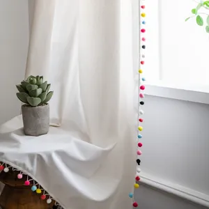 Detalle de cortina de lino crema con flecos de pompones de colores y luz natural suave