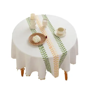 Mantel redondo beige con bordado de hojas y flecos sobre una mesa de madera