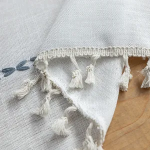 Macro detail of beige cotton linen fabric with blue dragonfly embroidery and tassels